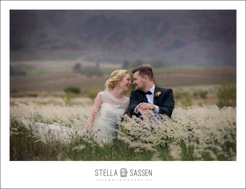  bride and groom laughing together sitting in a field of wild white flowers, soft mountains in the background, during an intimate wedding in the Cape Winelands.